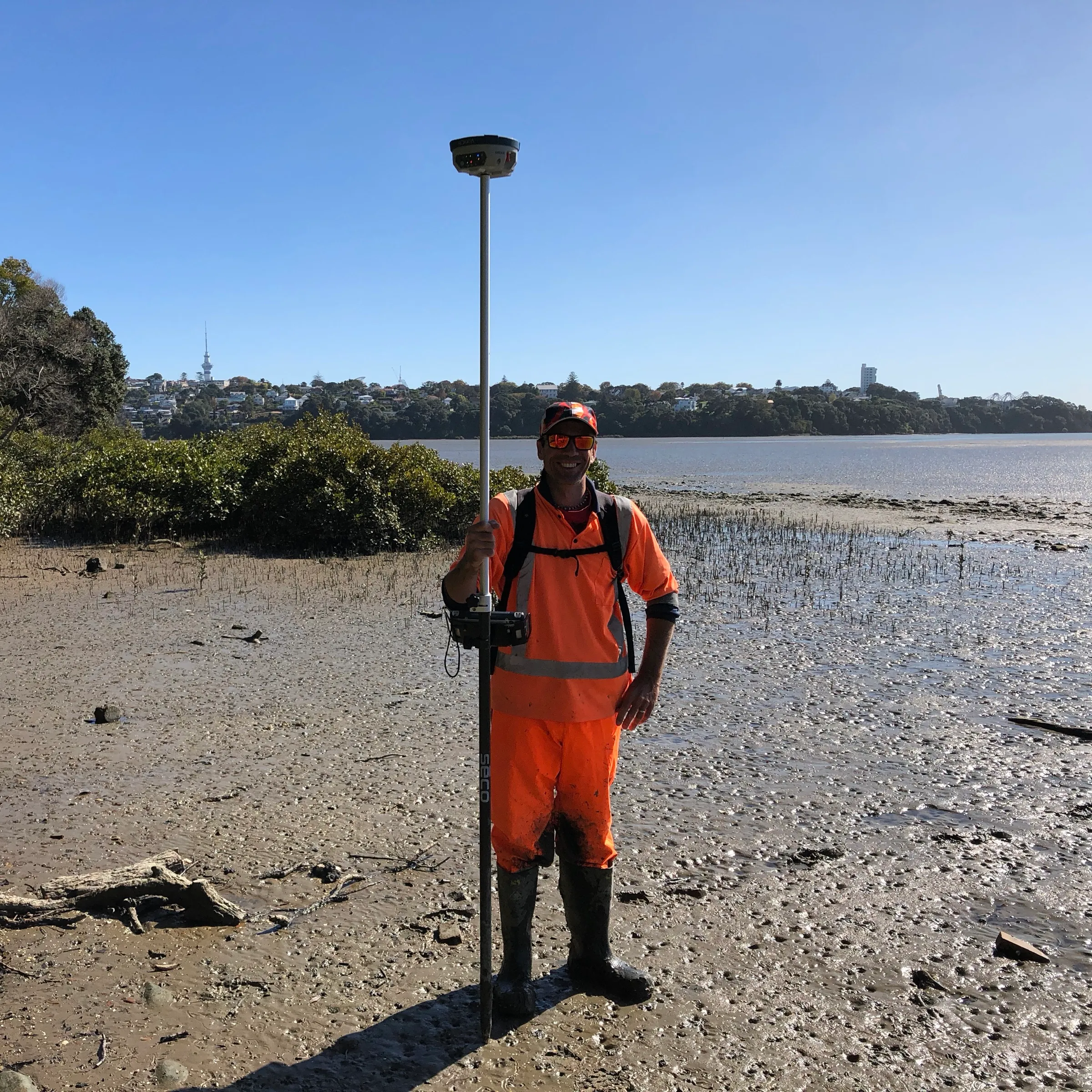 A surveyor with a GNSS pole on the mudflats at low tide, with the city skyline behind